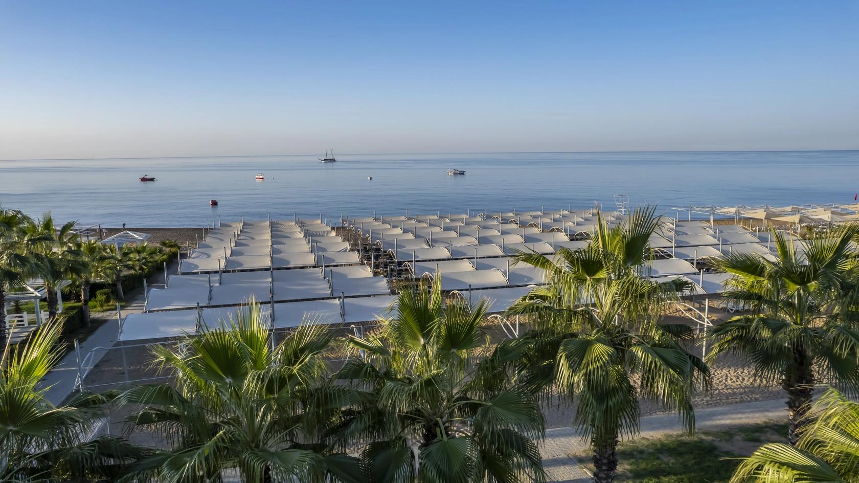 Strand med rækker af hvide parasoller og palmer, med både på det rolige blå hav. J´adore Deluxe Hotel og Spa, Side