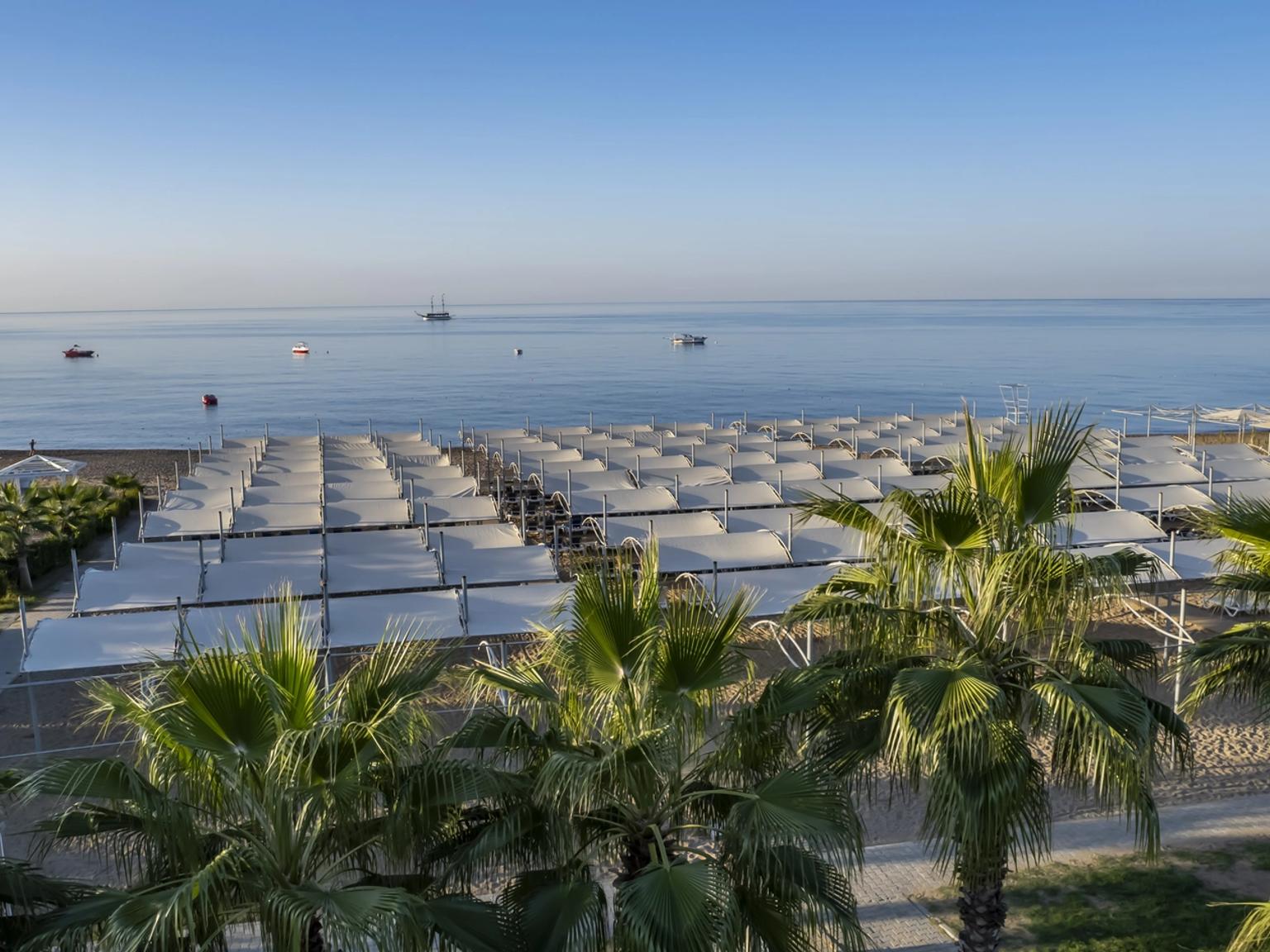 Strand med rækker af hvide parasoller og palmer, med både på det rolige blå hav. J´adore Deluxe Hotel og Spa, Side