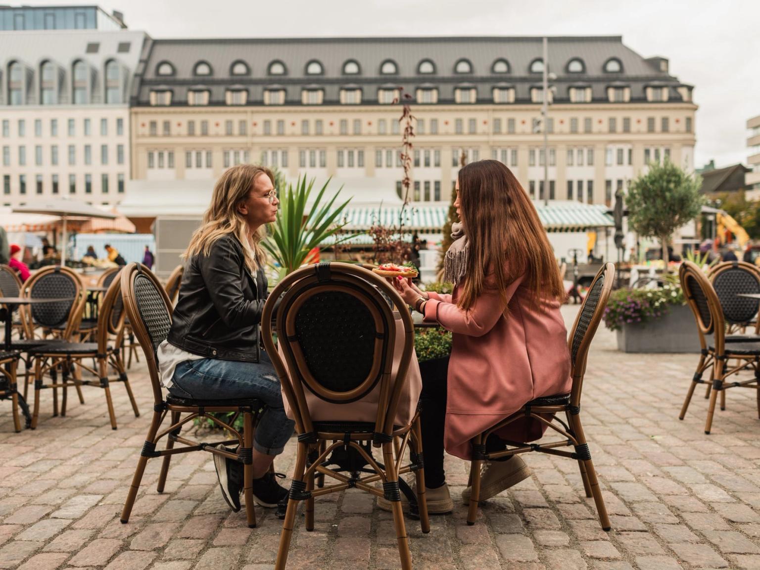 To kvinder snakker ved et udendørs cafébord på en byplads.