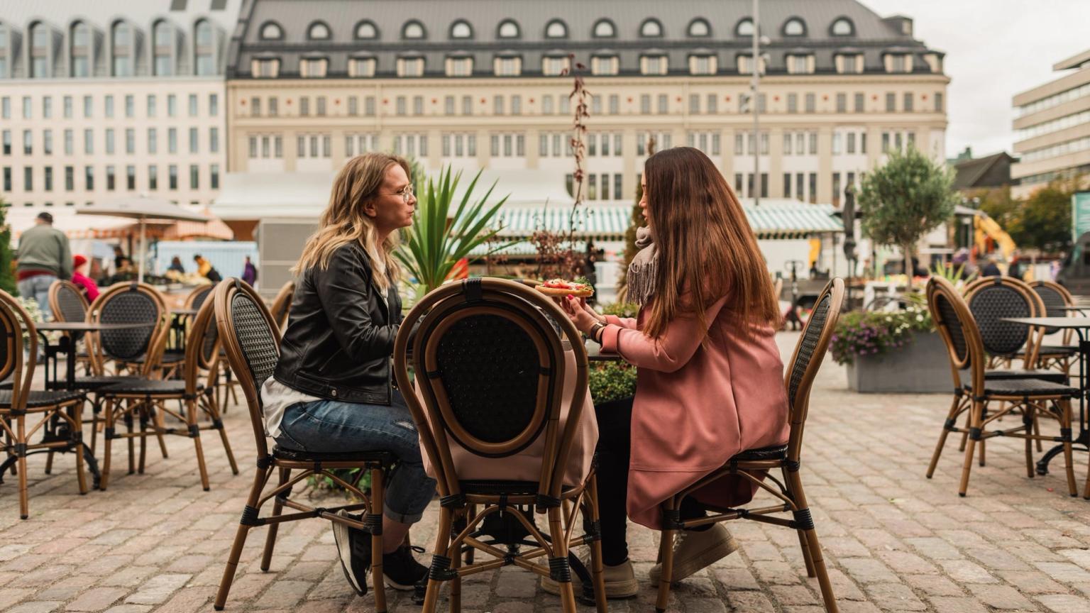 To kvinder sidder og taler ved et udendørs cafébord på en byplads.