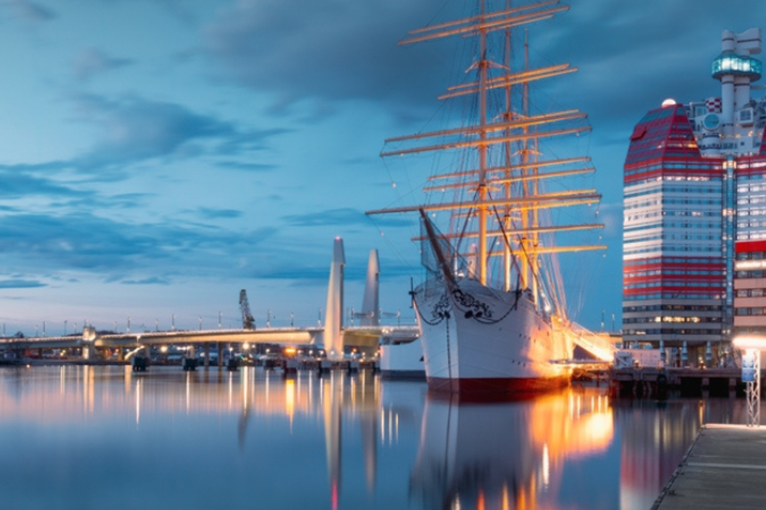 Stort sejlskib fortøjet i en byhavn i skumringen, med en rød og hvid skyskraber og en bro, der spejler sig i vandet.