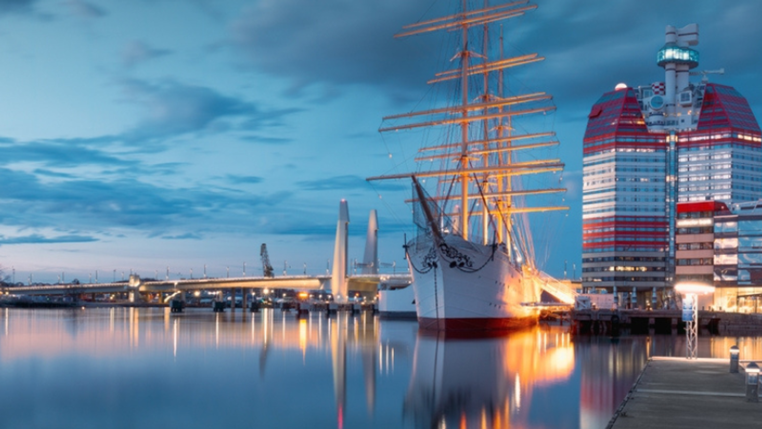 Stort sejlskib fortøjet i en byhavn i skumringen, med en rød og hvid skyskraber og en bro, der spejler sig i vandet.