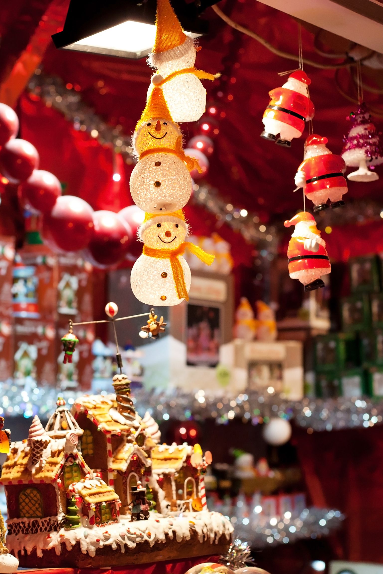 En starkt upplyst julutställning med hängande snögubbe- och tomteprydnader, ett pepparkakshus och en tomtefigur bland rött tyg och glitter.