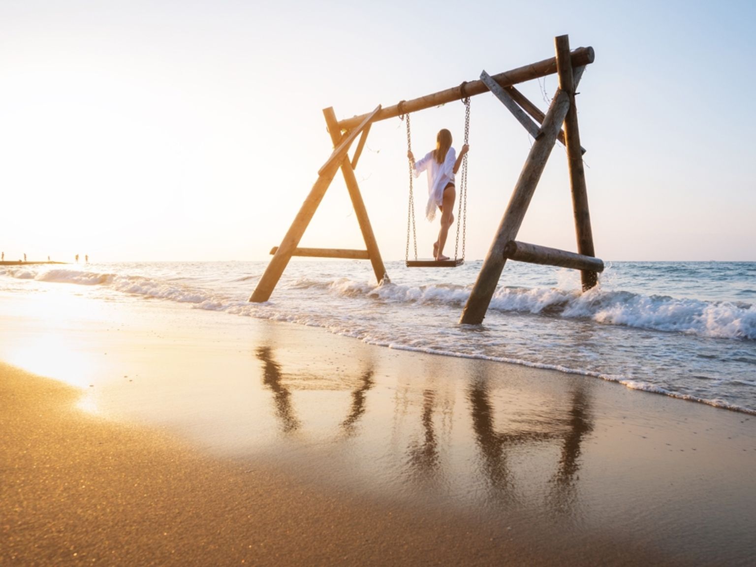 En kvinna gungar på en trägungställning på stranden ute i havet vid solnedgången.