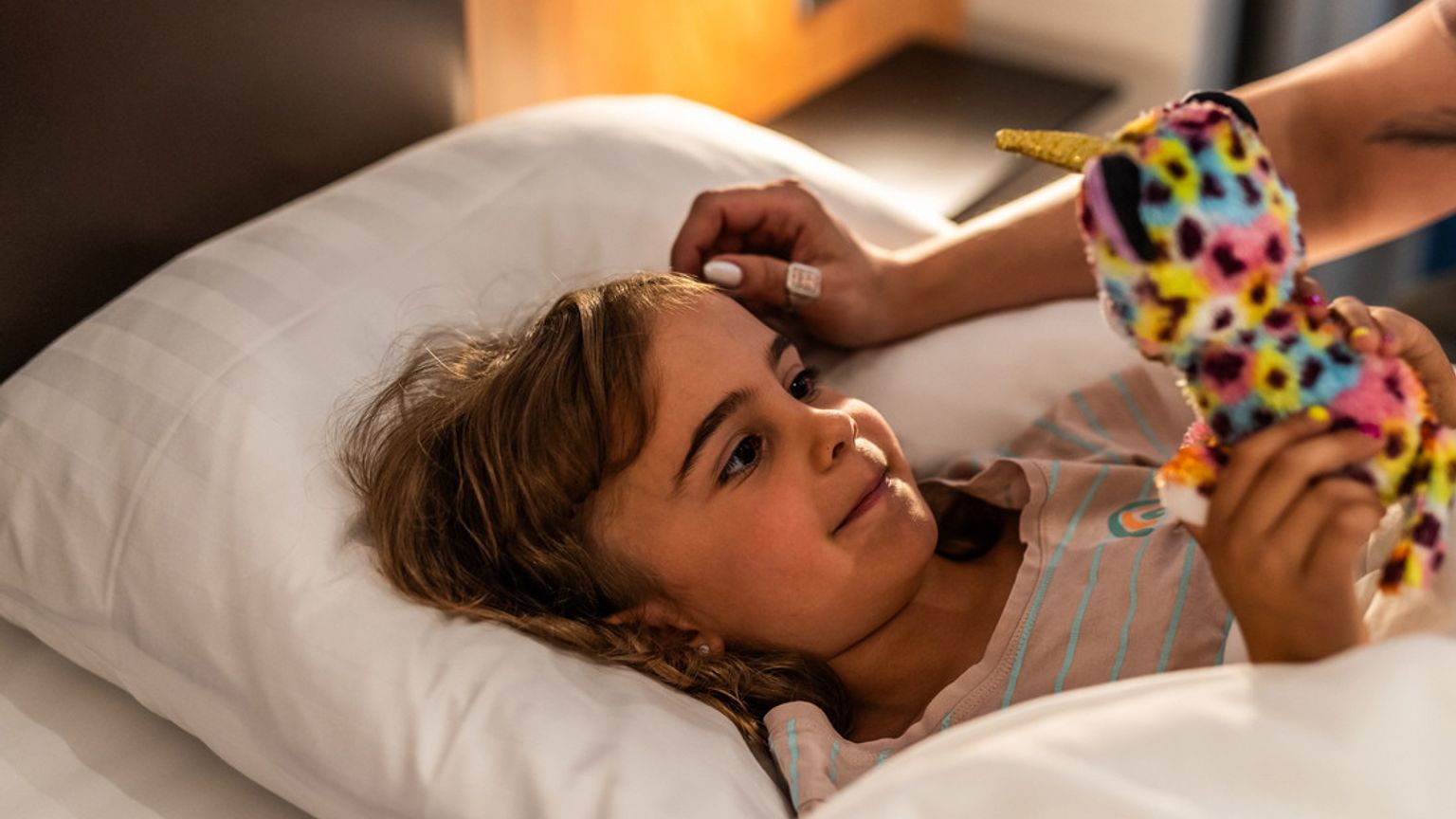 En leende ung flicka ligger i sängen och tittar på ett färgglatt enhörningsgosedjur. Billund Airport hotell