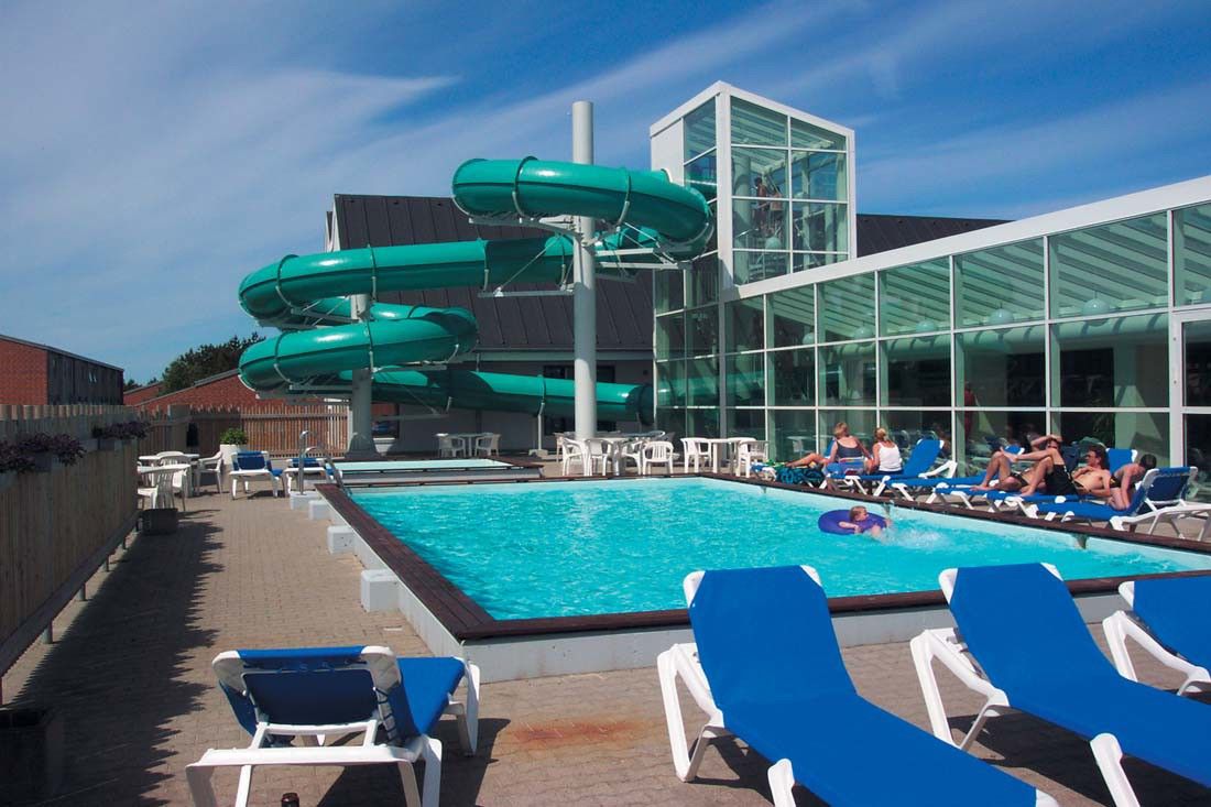 Et utendørs bassengområde med en svingete grønn vannsklie, solbadere og en moderne bygning med glassfasade. Landal Grønhøj Strand