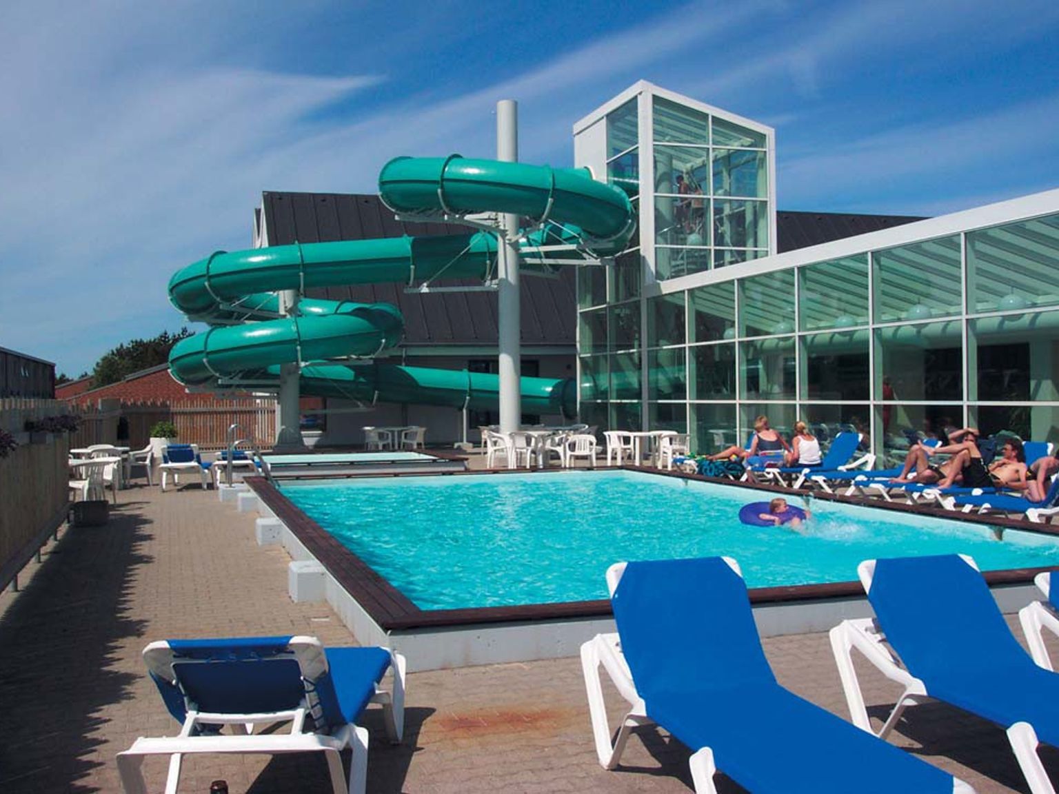 Et utendørs bassengområde med en svingete grønn vannsklie, solbadere og en moderne bygning med glassfasade. Landal Grønhøj Strand