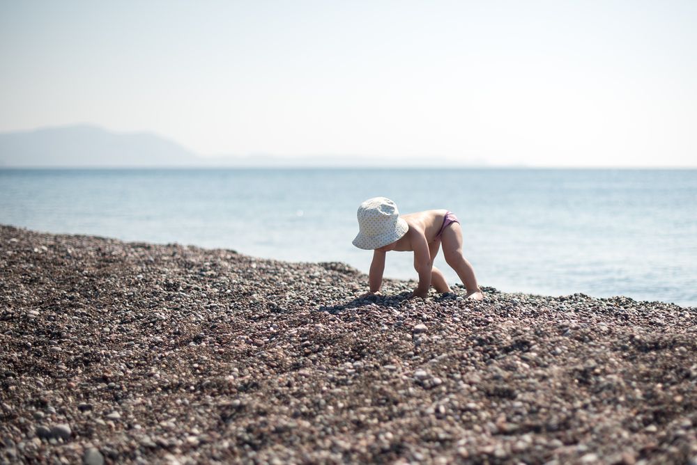 En baby med solhatt kryper på en steinete strand ved havet.