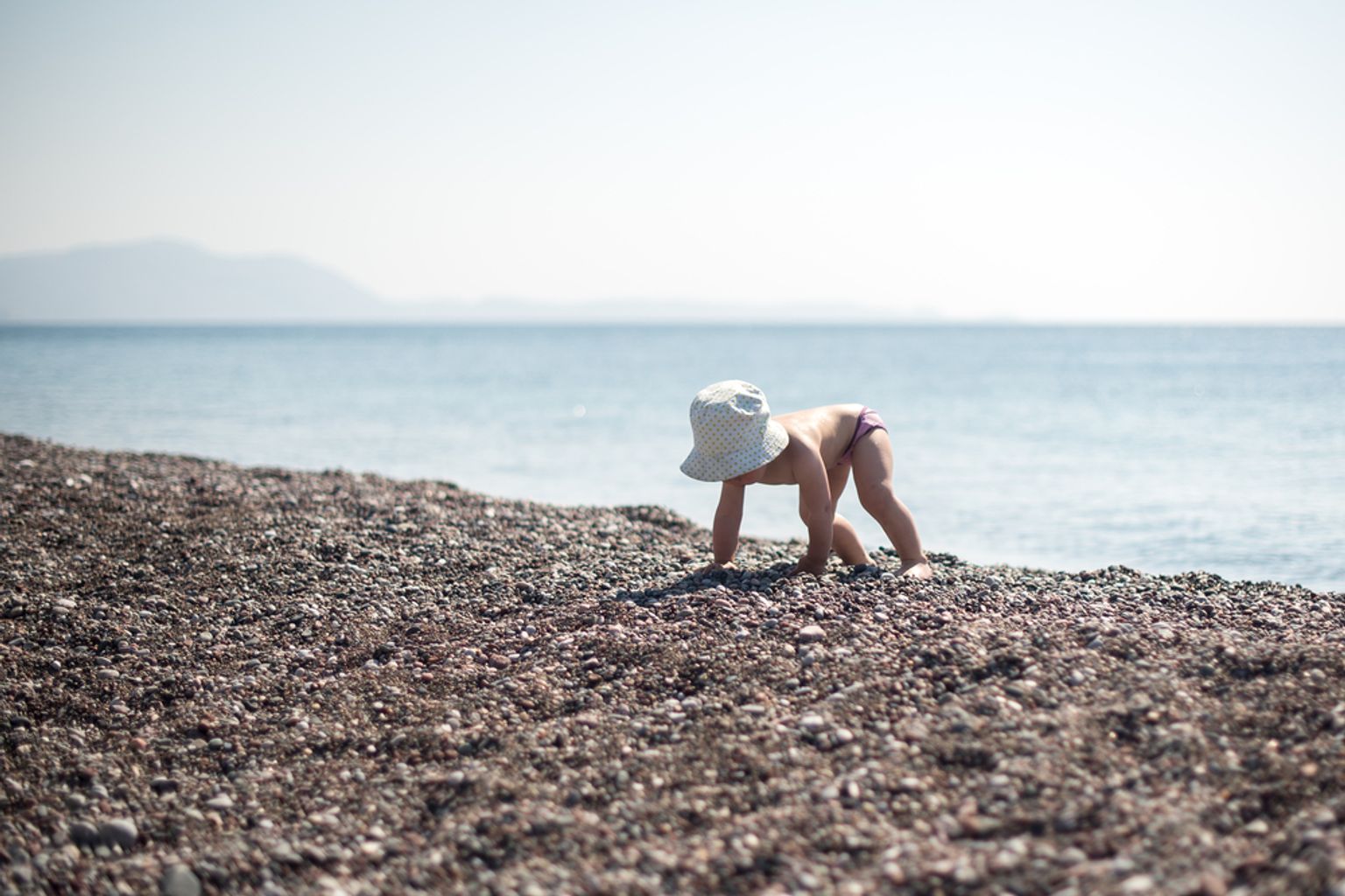 Een baby met een zonnehoed kruipt op een rotsachtig strand bij de oceaan.