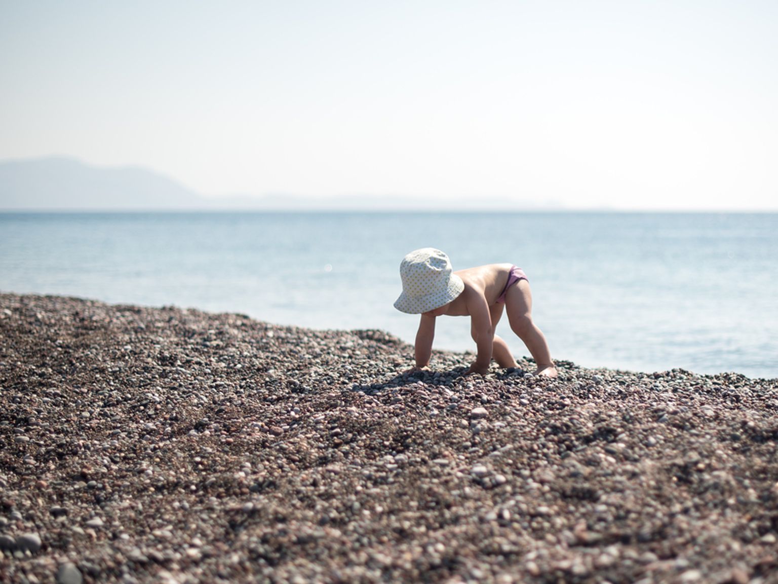 En baby med solhatt kryper på en steinete strand ved havet.