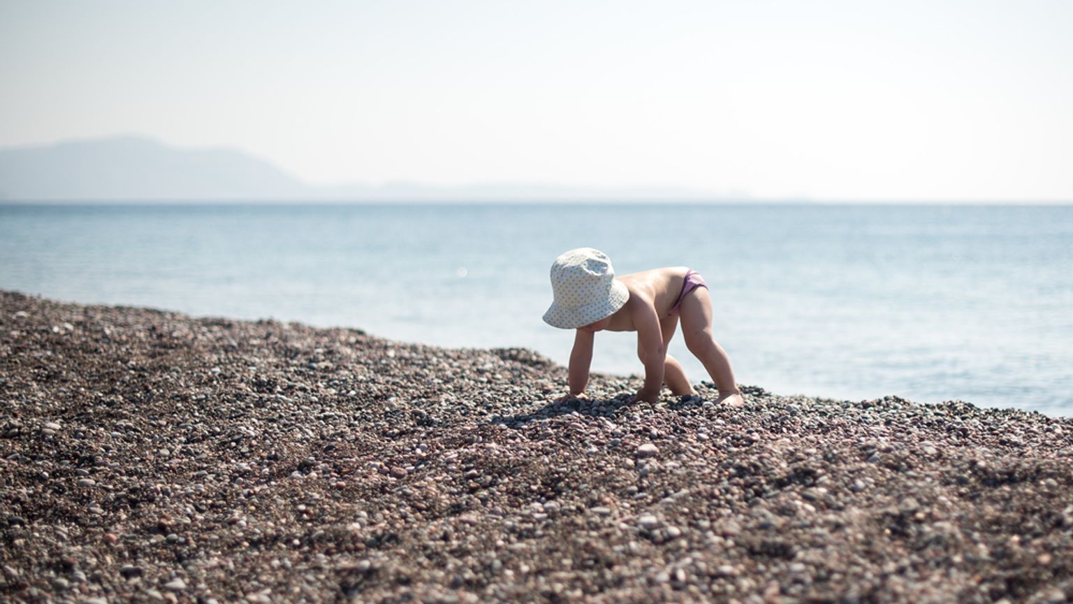 Een baby met een zonnehoed kruipt op een rotsachtig strand bij de oceaan.