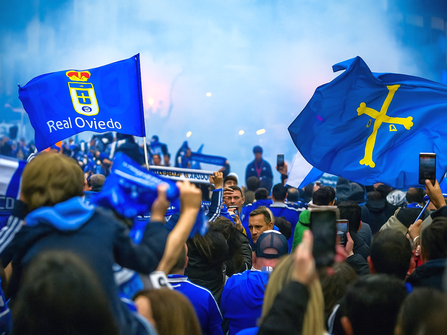 Eine große Menge von Real Oviedo-Fans feiert, schwenkt blaue Fahnen und Schals inmitten von Rauch.