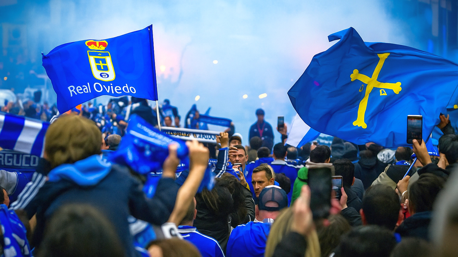 Een grote menigte Real Oviedo-fans viert feest, zwaaiend met blauwe vlaggen en sjaals te midden van rook.