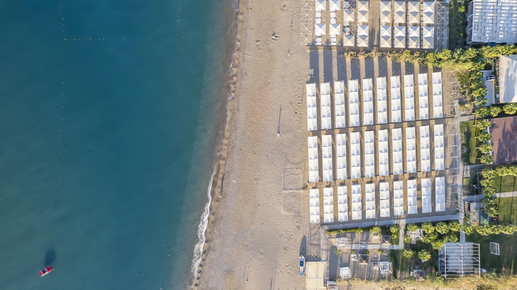 Luftfoto af en sandstrand med rækker af hvide cabanas, ved siden af et blåt hav med en lille rød båd. J'adore Deluxe Hotel og Spa, Side