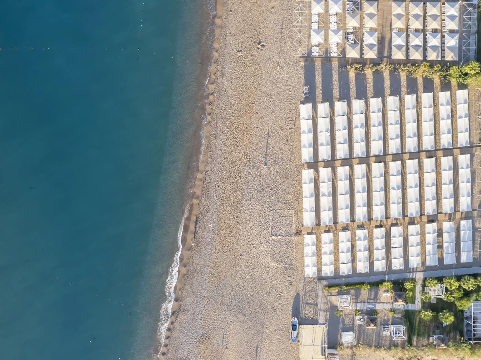 Luftfoto af en sandstrand med rækker af hvide cabanas, ved siden af et blåt hav med en lille rød båd. J'adore Deluxe Hotel og Spa, Side