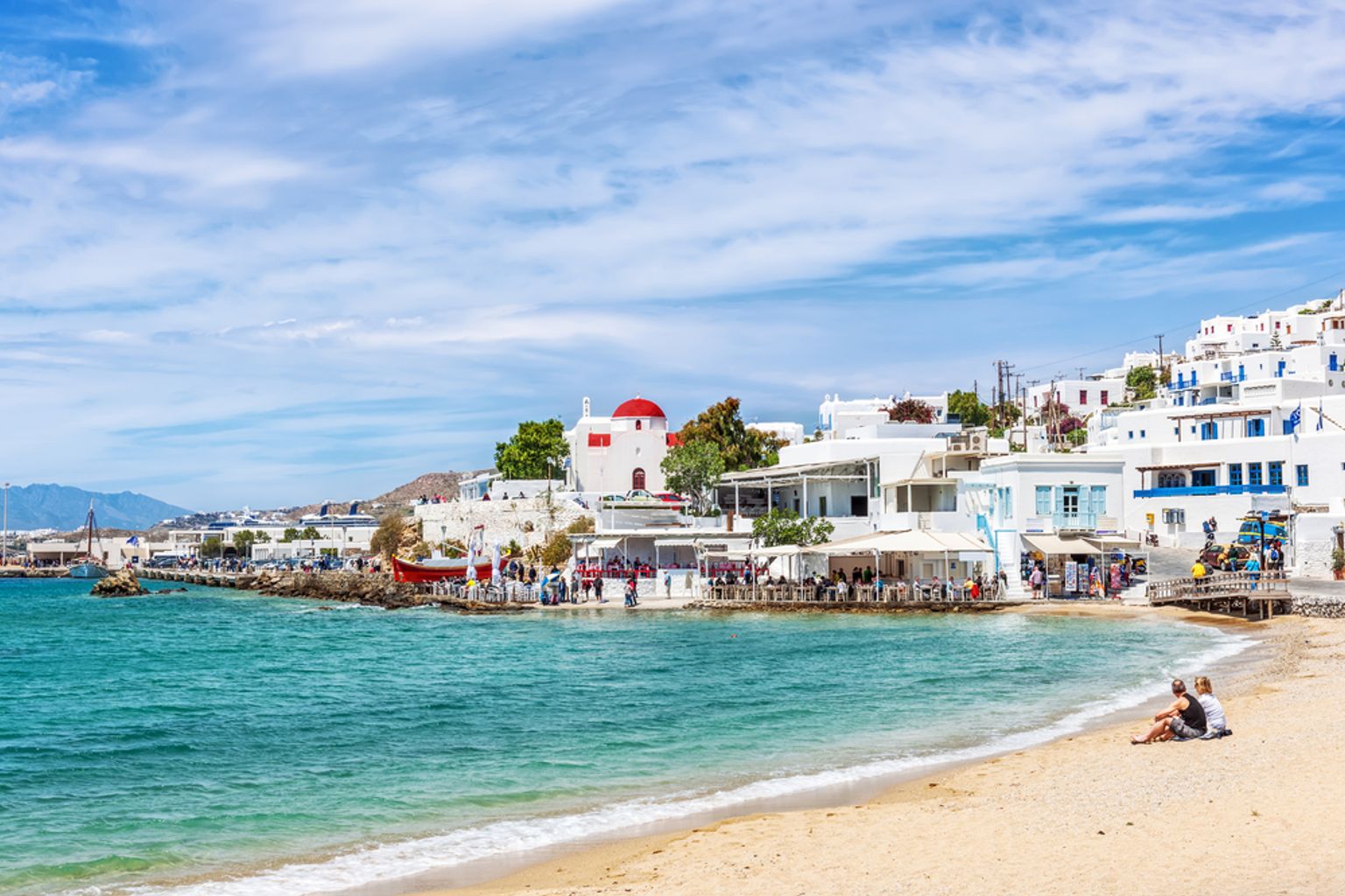 Greek island town of white buildings and a red-domed church lining a turquoise bay with a sandy beach. Mykonos, Greece