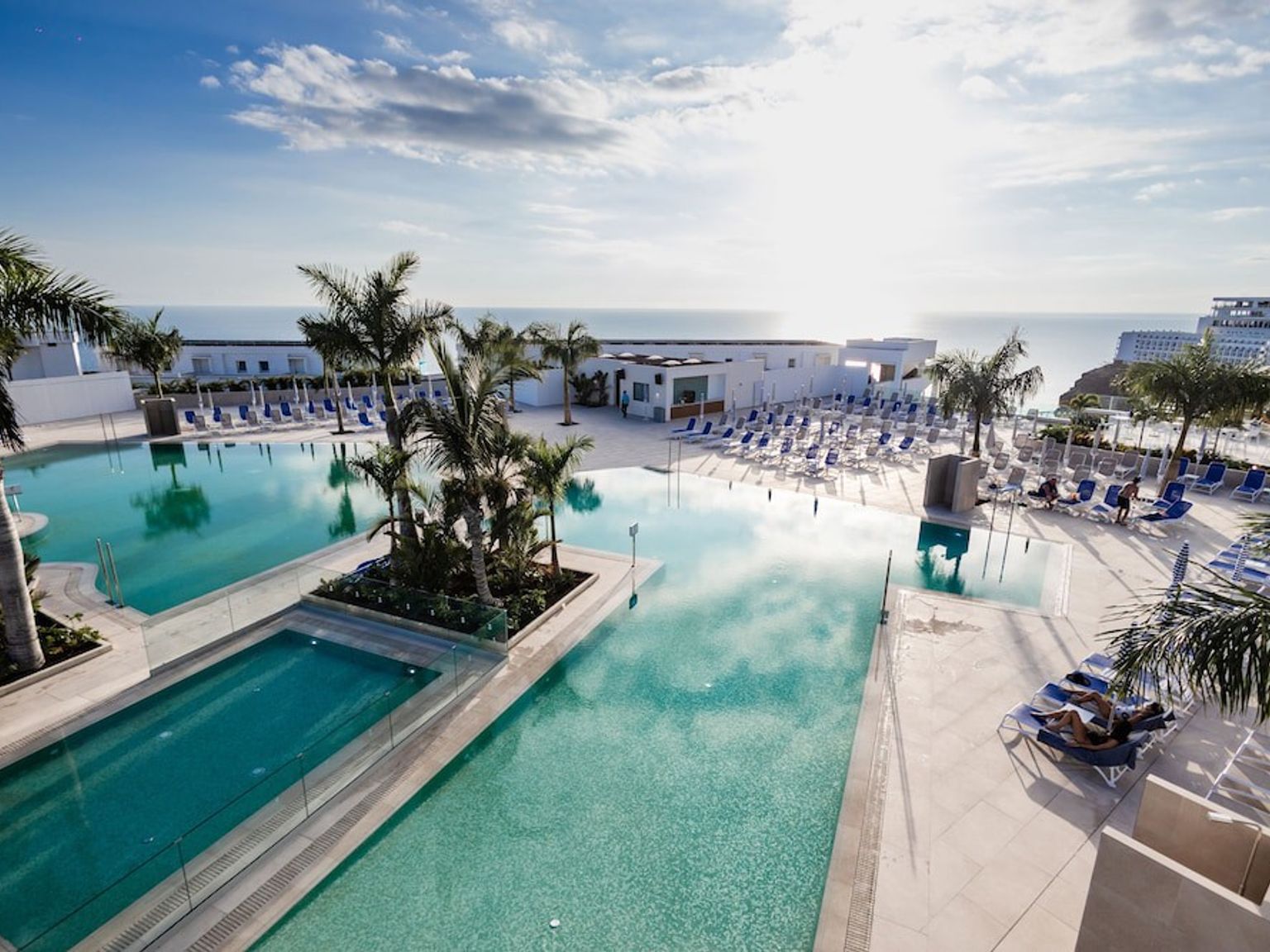 Oversigt over en solrig resort med flere swimmingpools, palmer og hav i baggrunden.