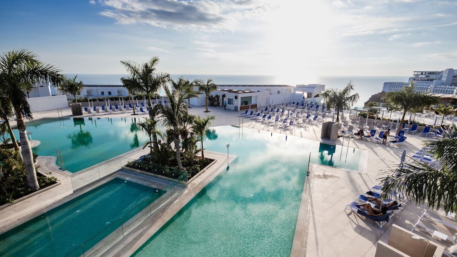 Oversigt over en solrig resort med flere swimmingpools, palmer og hav i baggrunden.