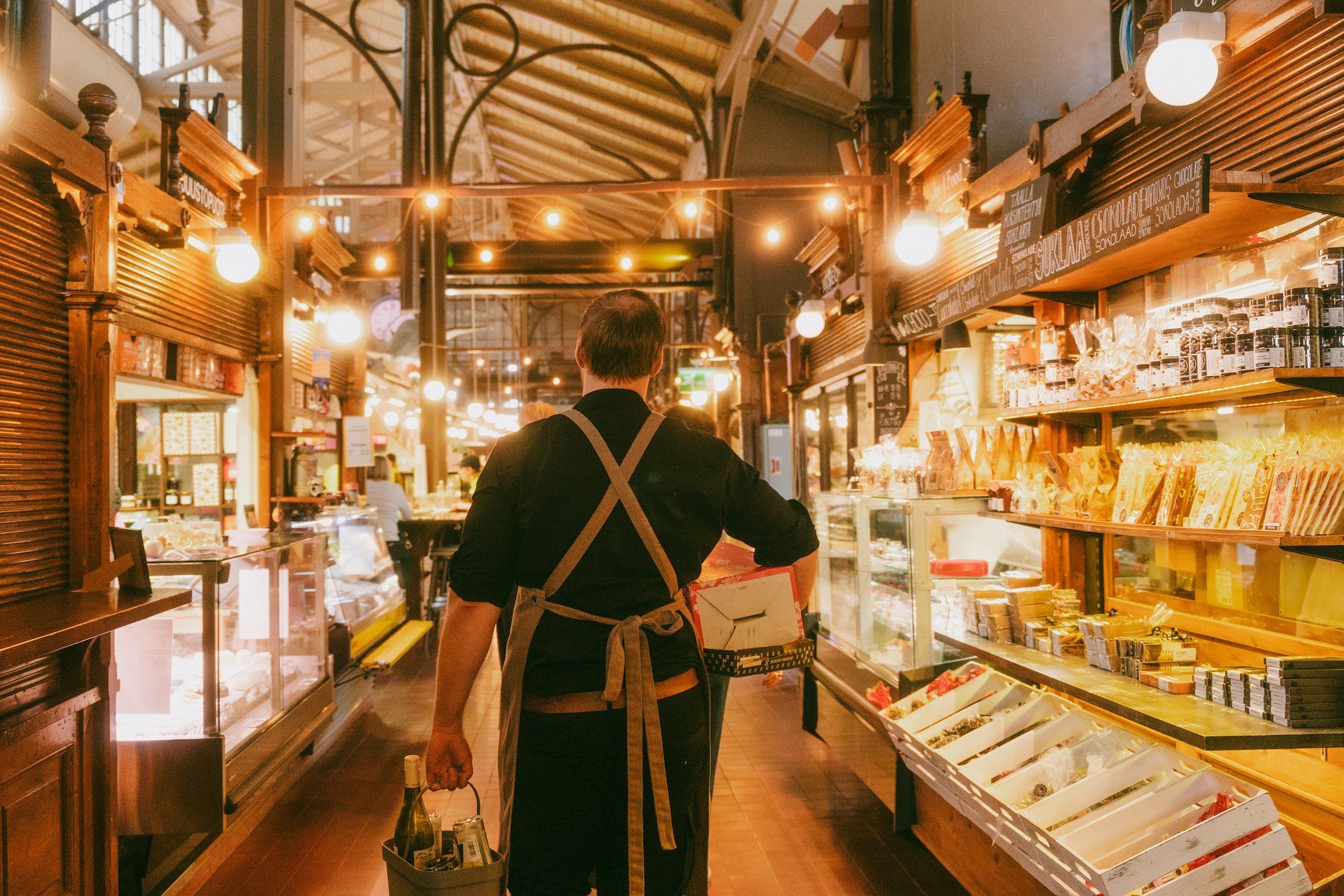 En mand i et forklæde går gennem en varmt oplyst markedsbygning og bærer en kasse og vinflasker forbi madboder.