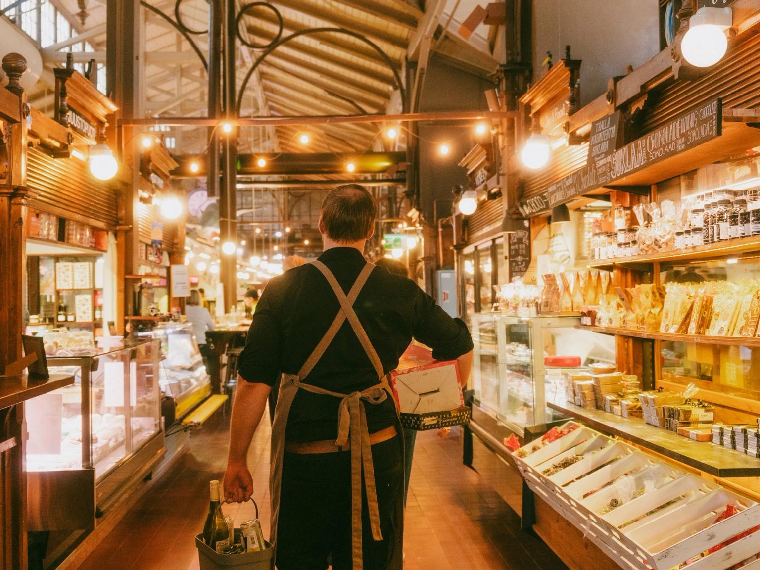 En mand i et forklæde går gennem en varmt oplyst markedsbygning og bærer en kasse og vinflasker forbi madboder.