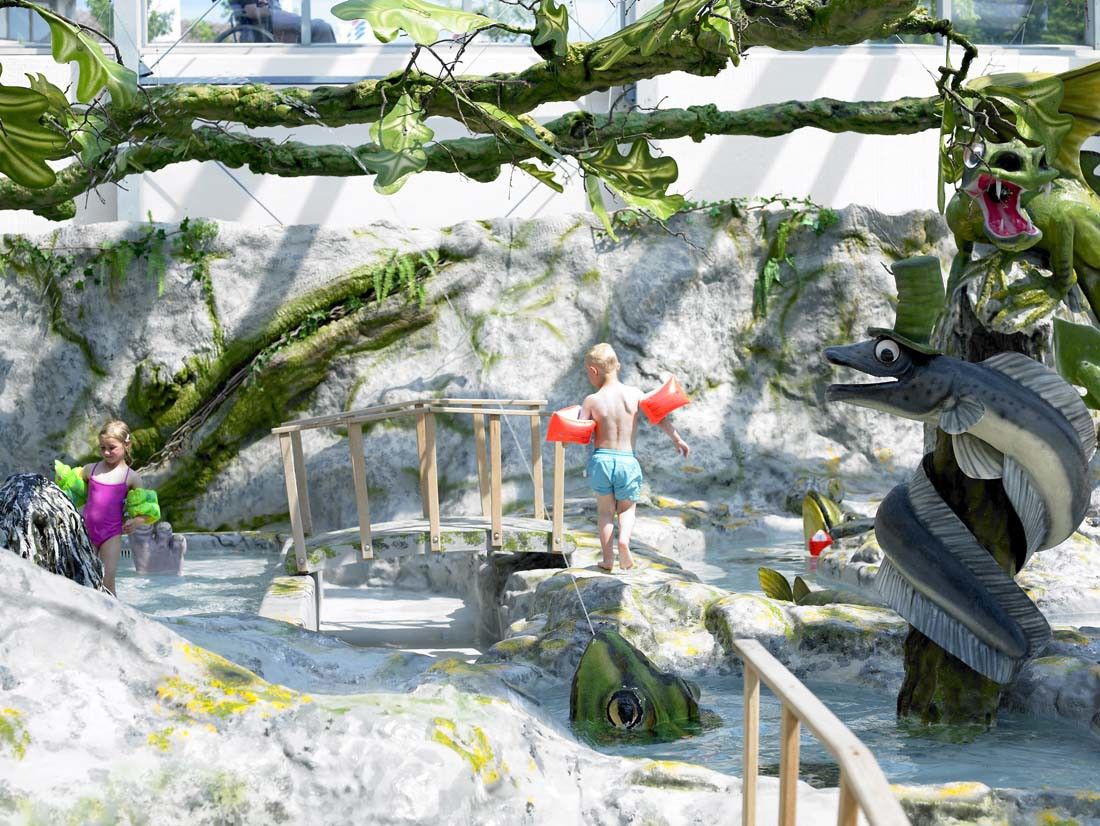 Barn leker i en fantasifull vannpark med steinformasjoner, kunstige trær og store statuer av en drage og en fisk med flosshatt.