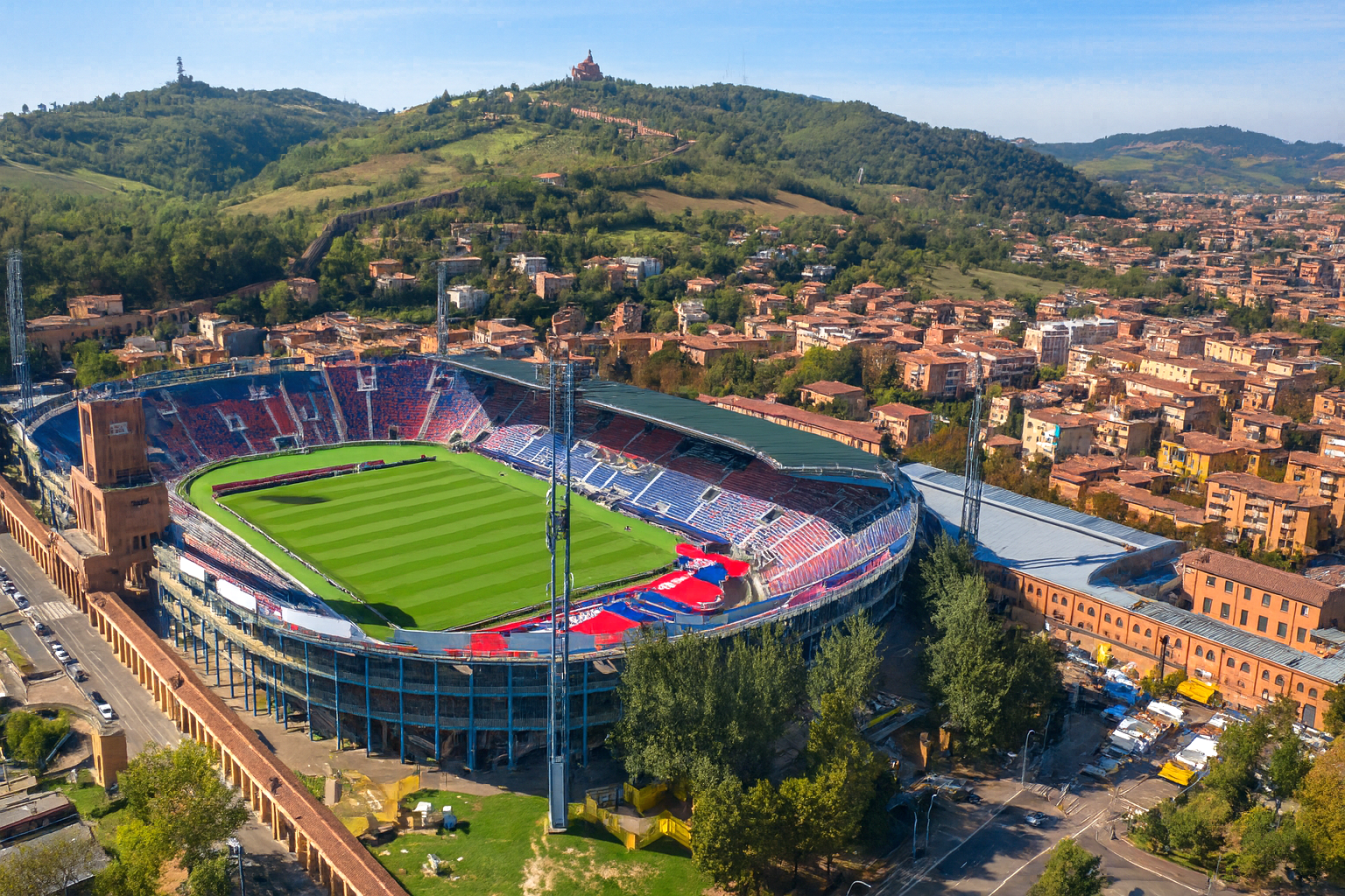 Aerial view of a large sports stadium with a green field, surrounded by a city and hills.