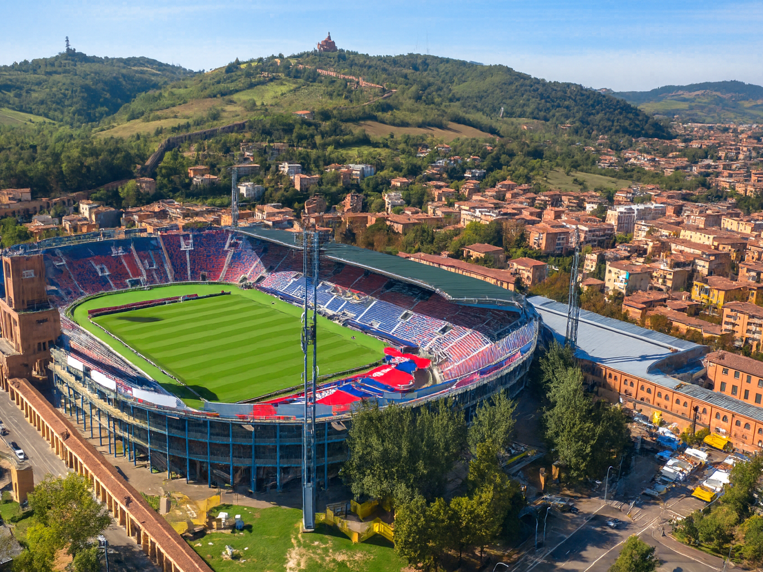 Légi felvétel egy nagy sportstadionról zöld pályával, város és dombok veszik körül.