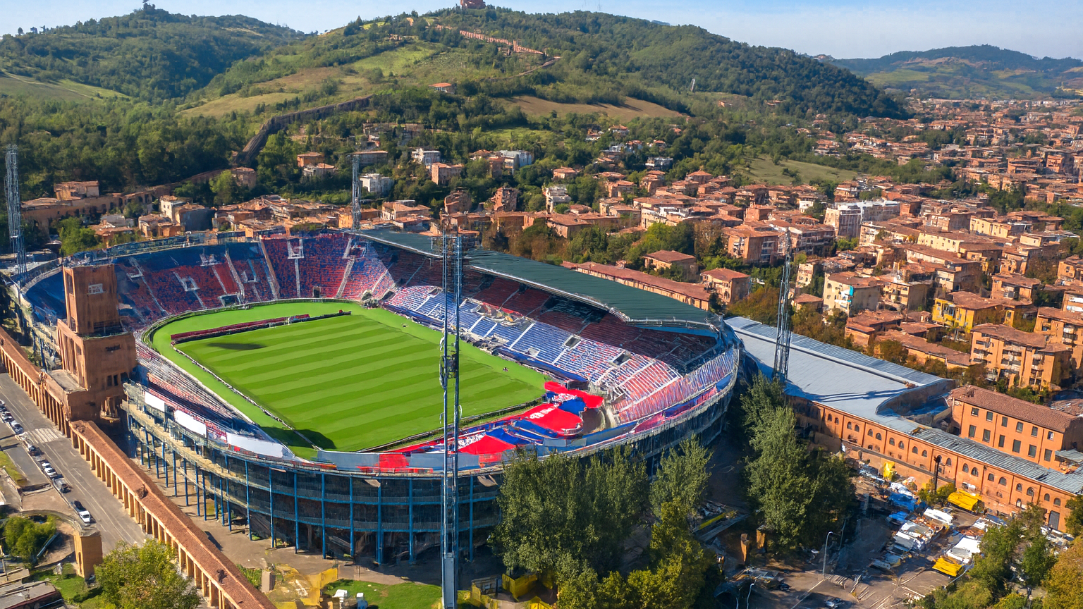 Aerial view of a large sports stadium with a green field, surrounded by a city and hills.