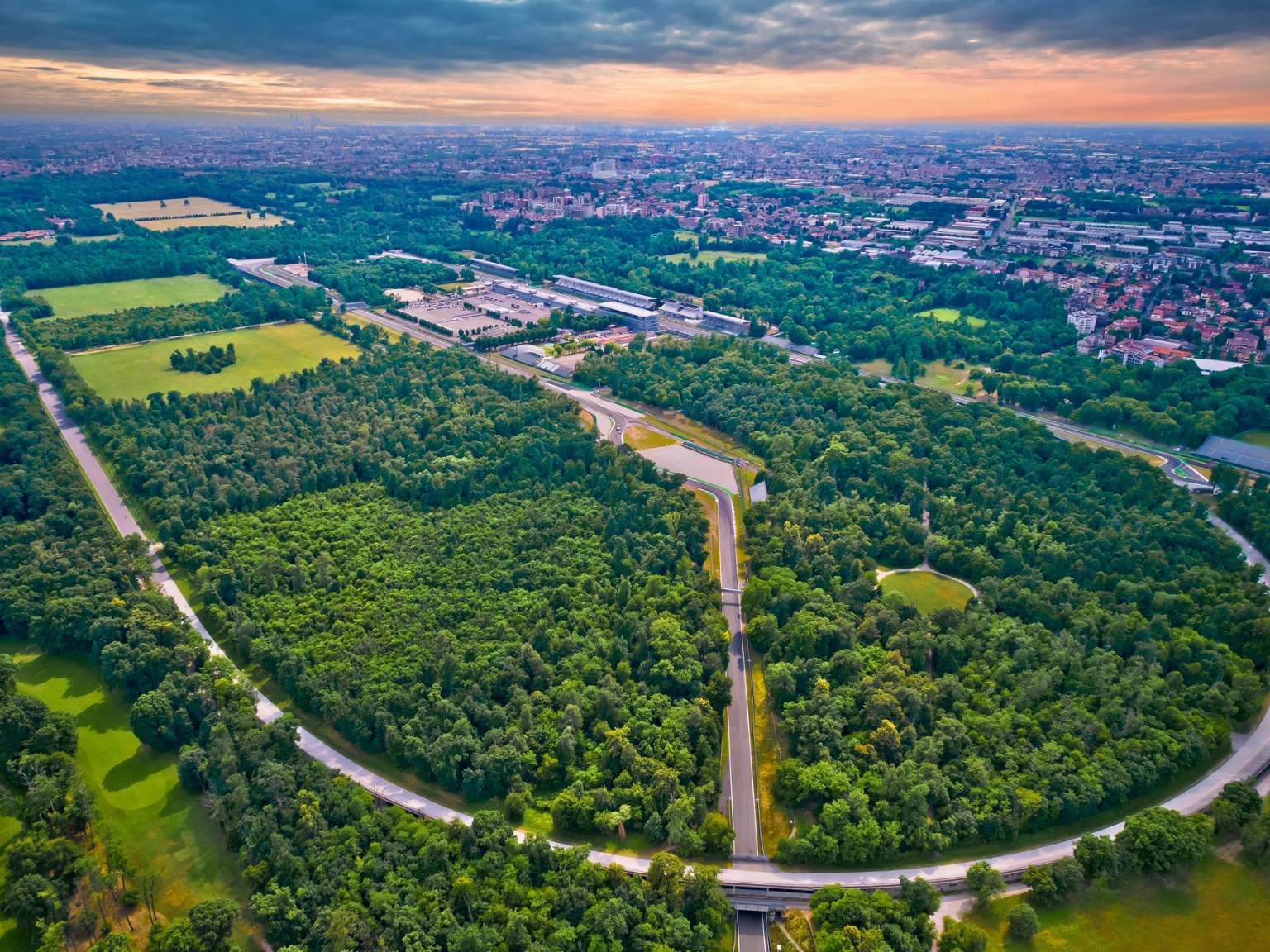 Widok z lotu ptaka na szeroką, krętą drogę biegnącą przez gęsty las, z miastem w tle.