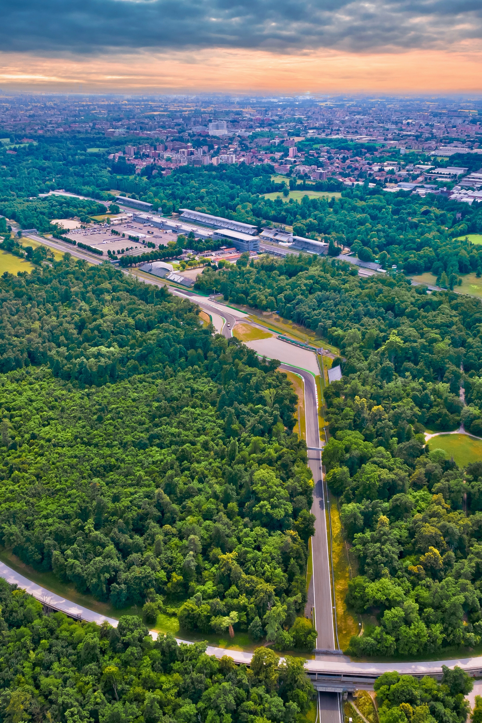 Widok z lotu ptaka na szeroką, krętą drogę biegnącą przez gęsty las, z miastem w tle.