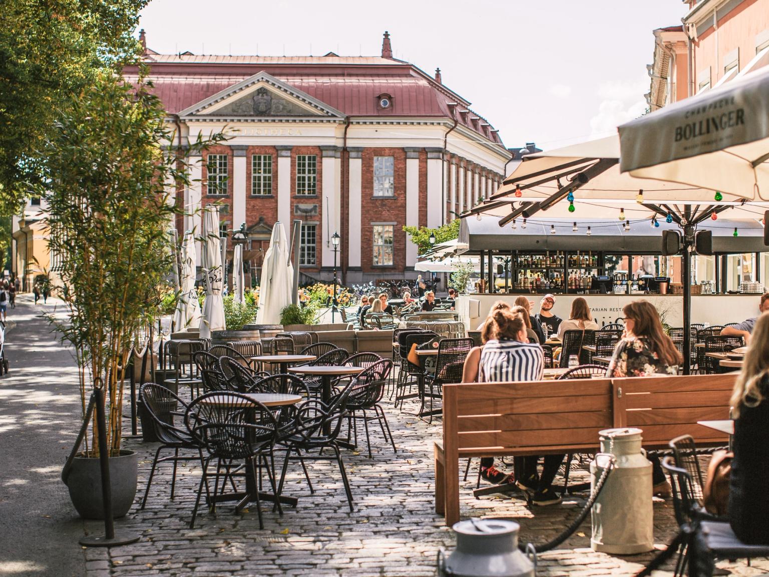Udendørs caféscene på en brostensbelagt gade med mennesker, der spiser, en storslået murstensbygning i baggrunden.