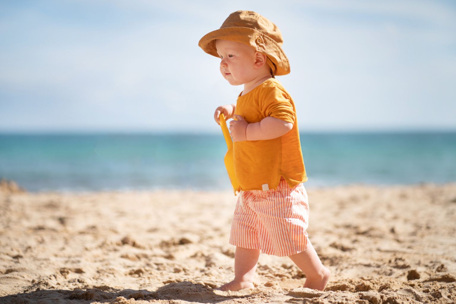 En bebis i solhatt och randiga shorts går på en sandstrand och håller i en gul leksaksspade, med havet i bakgrunden.