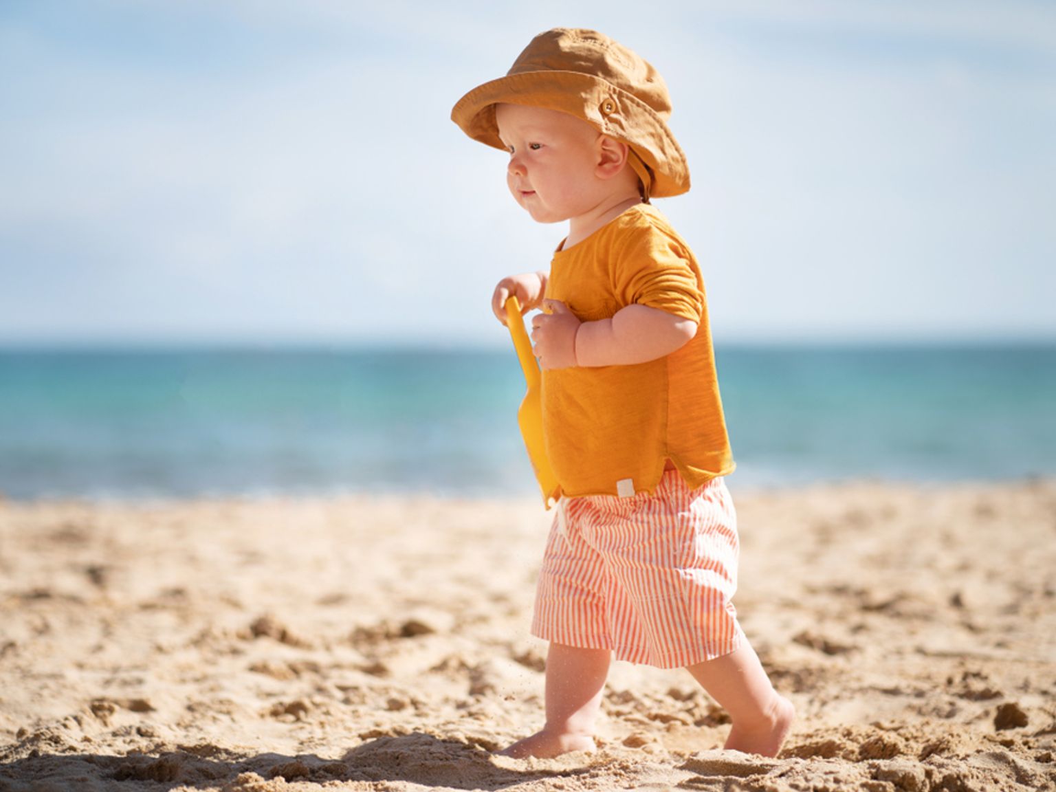En bebis i solhatt och randiga shorts går på en sandstrand och håller i en gul leksaksspade, med havet i bakgrunden.