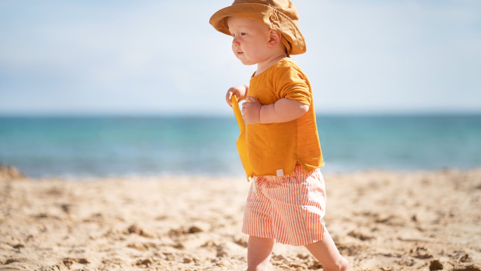 En bebis i solhatt och randiga shorts går på en sandstrand och håller i en gul leksaksspade, med havet i bakgrunden.