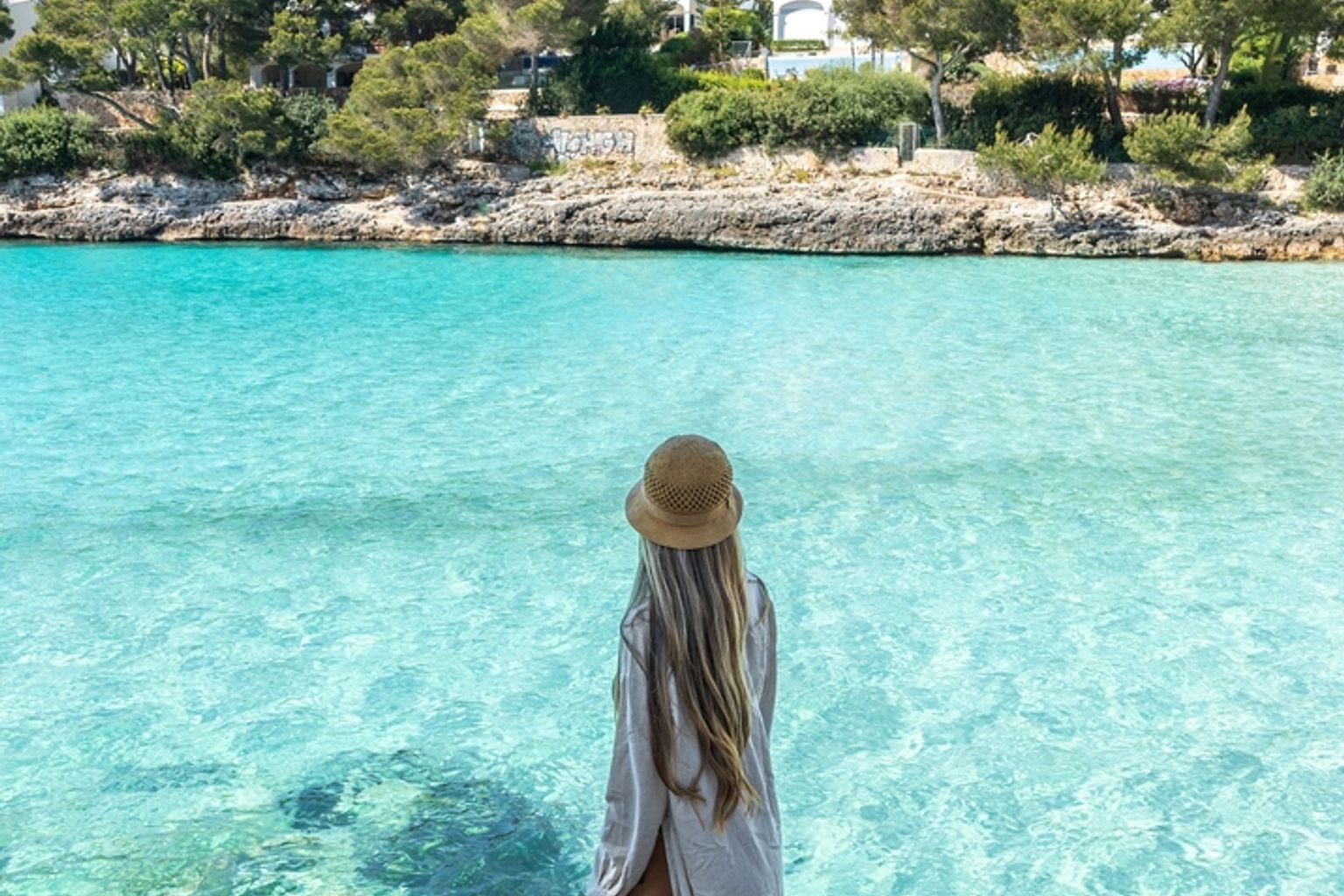 Kvinna på klippor med utsikt över en ljus turkos bukt med avlägsna byggnader och träd.
