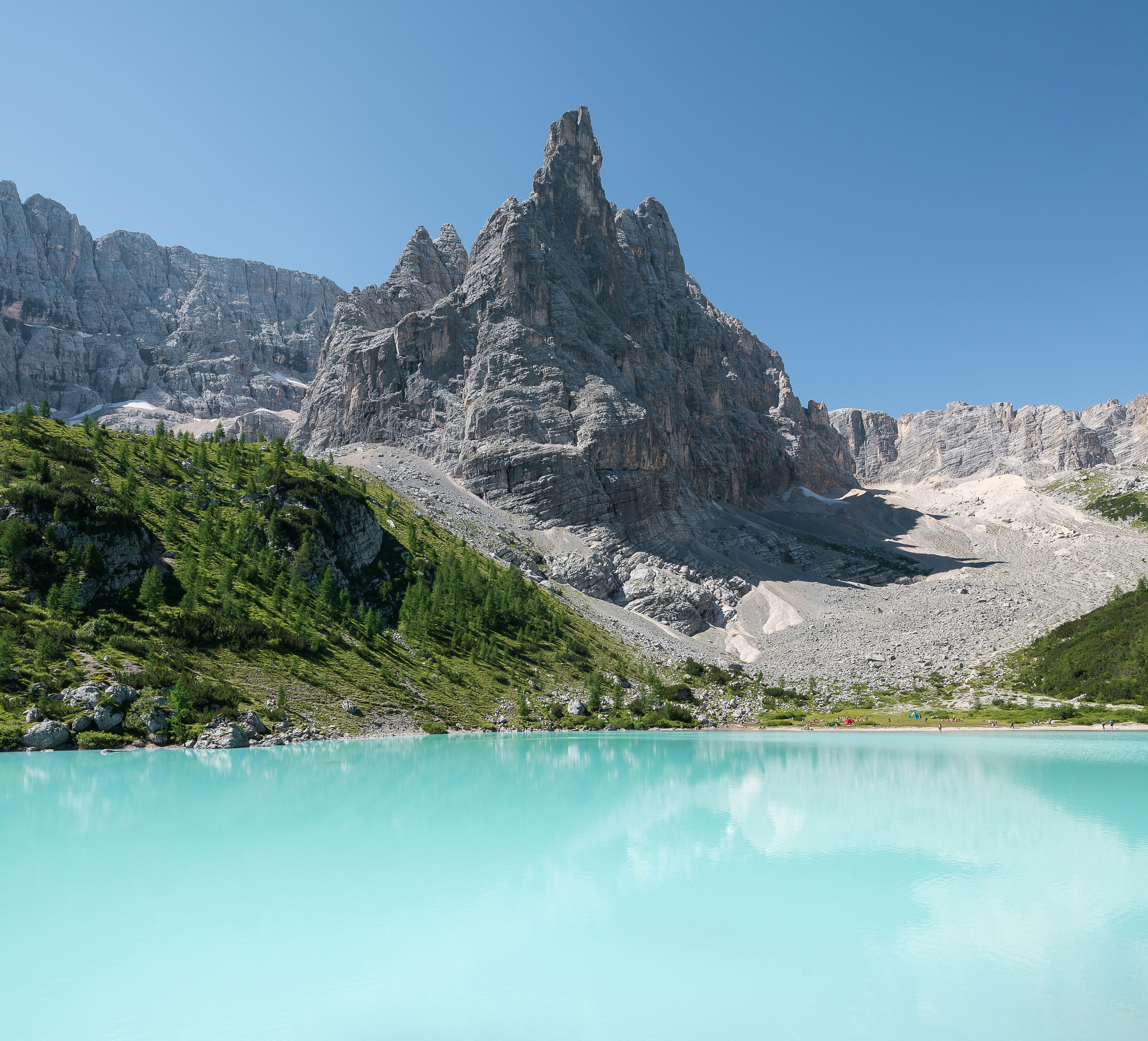 En livlig turkis innsjø speiler en klar blå himmel, med grønne skråninger og et høyt, taggete grått fjell bak den. Lago di Sorapis, Italia