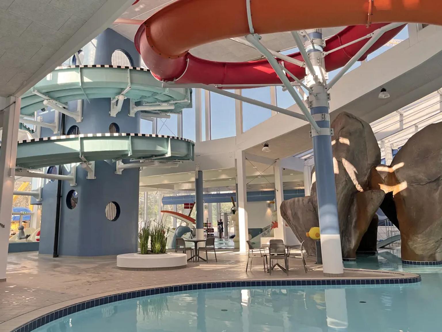 Innendørs vannpark med et blått sklietårn, rød og oransje rørsklie, svømmebasseng og steinfunksjoner.