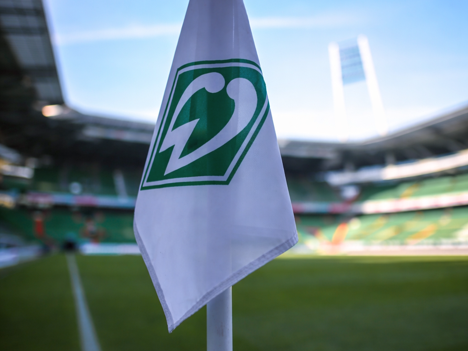 Bandera de esquina blanca con el logo verde de Werder Bremen en un campo de fútbol, estadio al fondo.