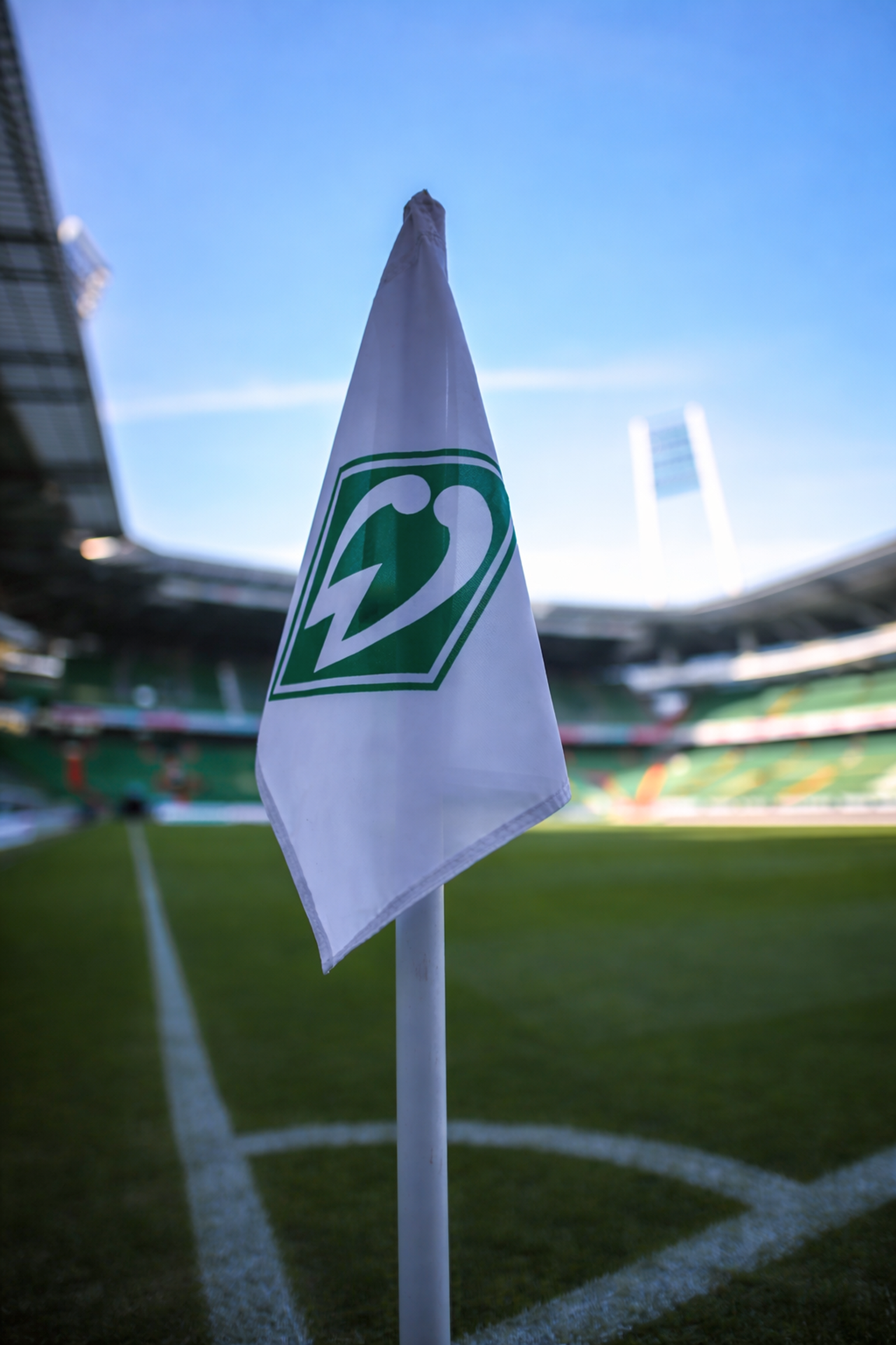 Bandera de esquina blanca con el logo verde de Werder Bremen en un campo de fútbol, estadio al fondo.