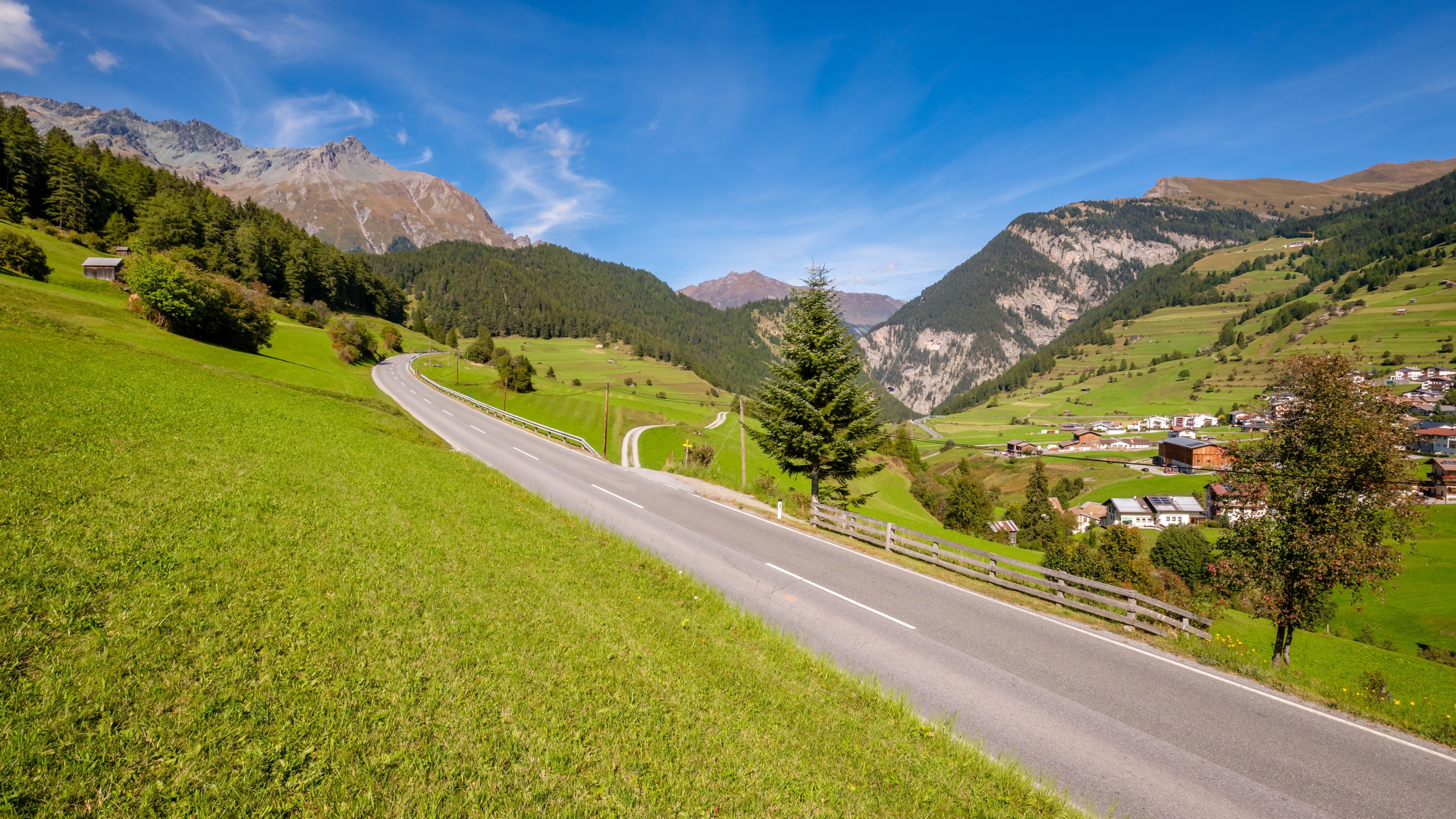 Fotografi på en landevej gennem grønne enge i Østrig med Alperne og hytter i baggrunden