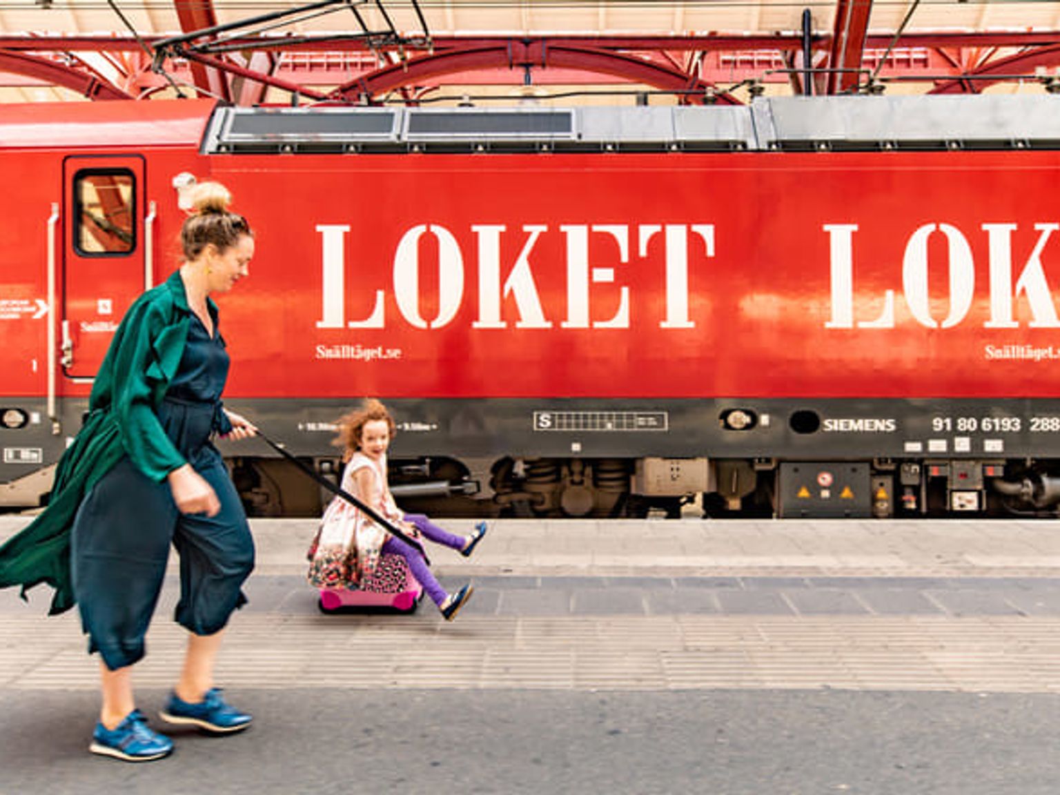 En kvinna drar ett barn på en rosa åkresväska längs en tågplattform bredvid ett rött tåg märkt "LOKET".