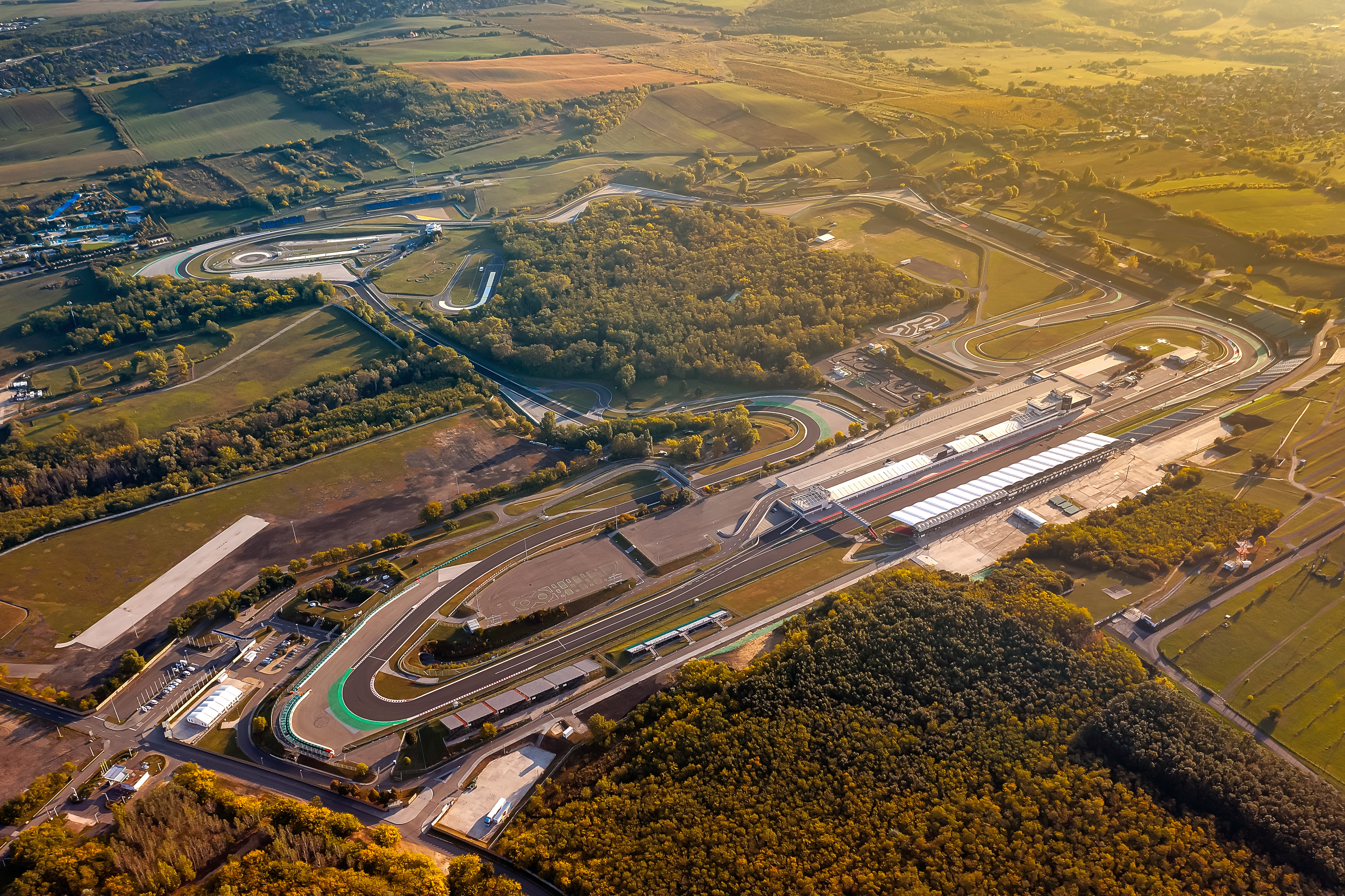Flyfoto av en racerbane med pitlane og tribuner, omgitt av grønt landskap i gyllent lys.