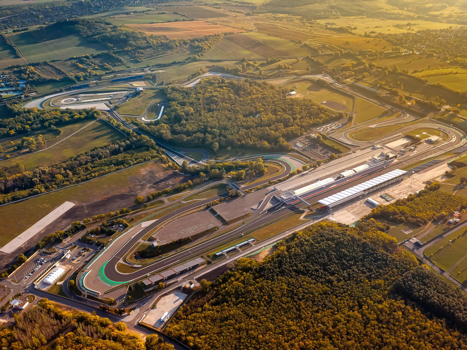 Flyfoto av en racerbane med pitlane og tribuner, omgitt av grønt landskap i gyllent lys.