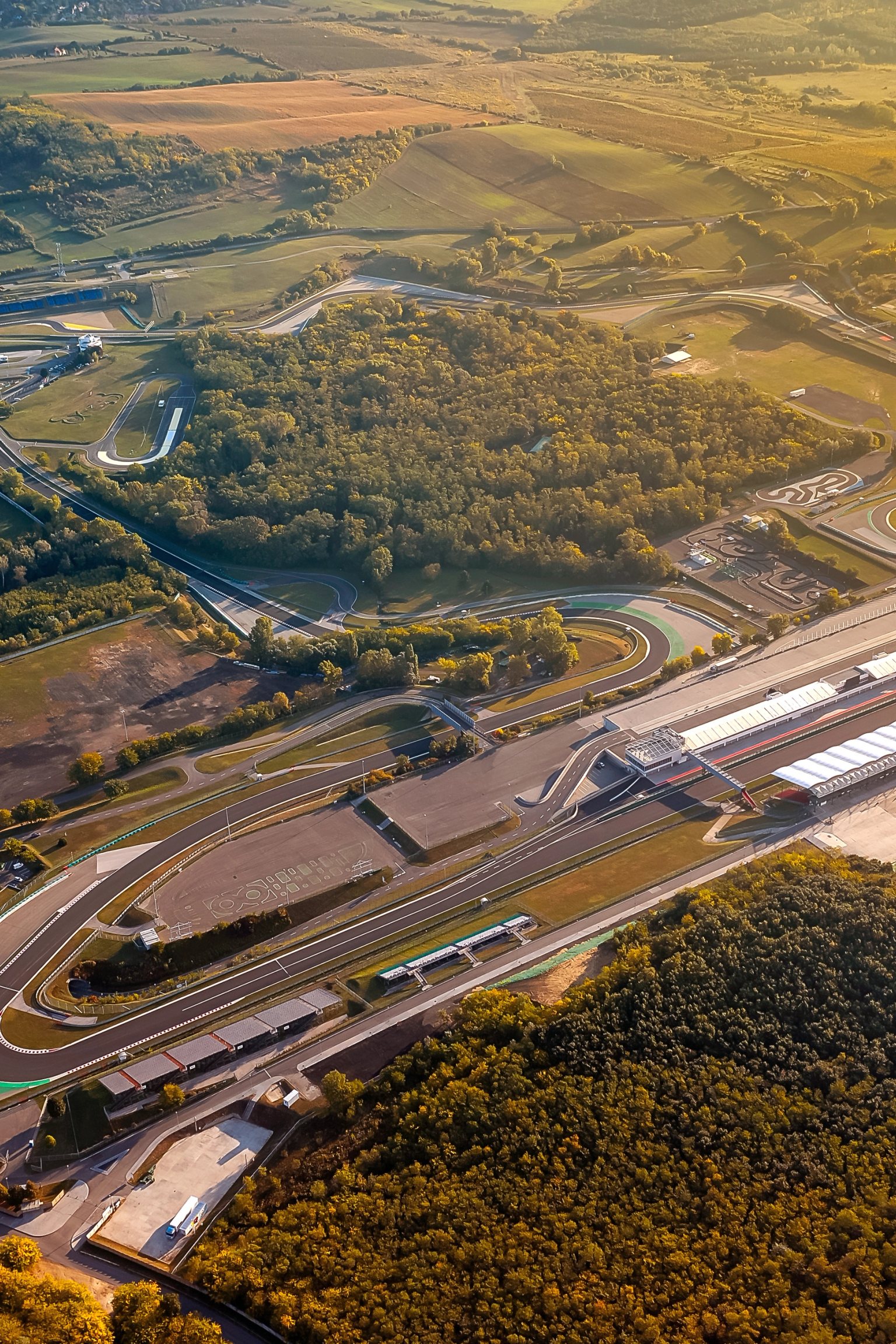 Flyfoto av en racerbane med pitlane og tribuner, omgitt av grønt landskap i gyllent lys.