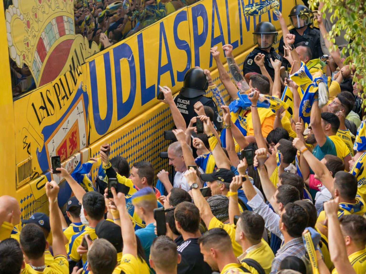 Una multitud densa de aficionados al fútbol animando en amarillo y azul rodea un autobús del equipo amarillo de UD Las Palmas, con policías presentes.