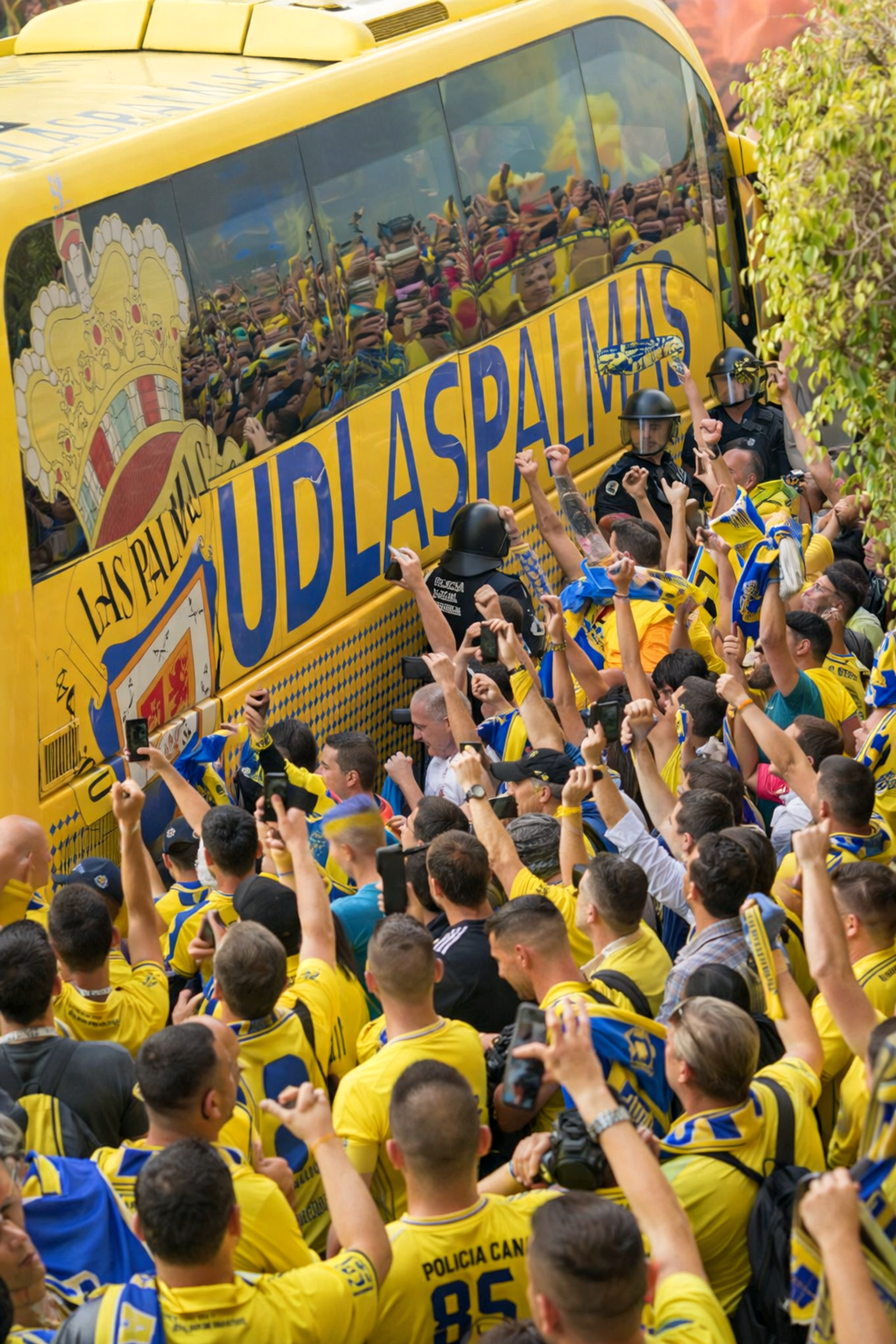 Una multitud densa de aficionados al fútbol animando en amarillo y azul rodea un autobús del equipo amarillo de UD Las Palmas, con policías presentes.
