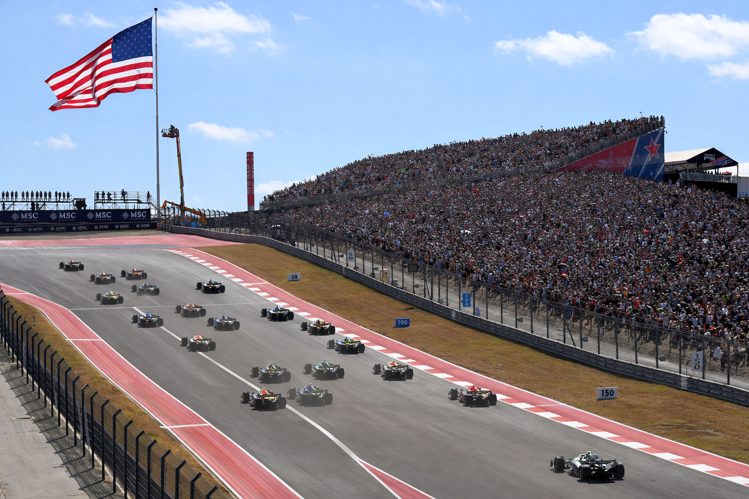 Open-wheel race cars on a track with a massive crowd in the grandstand, under a large American flag.