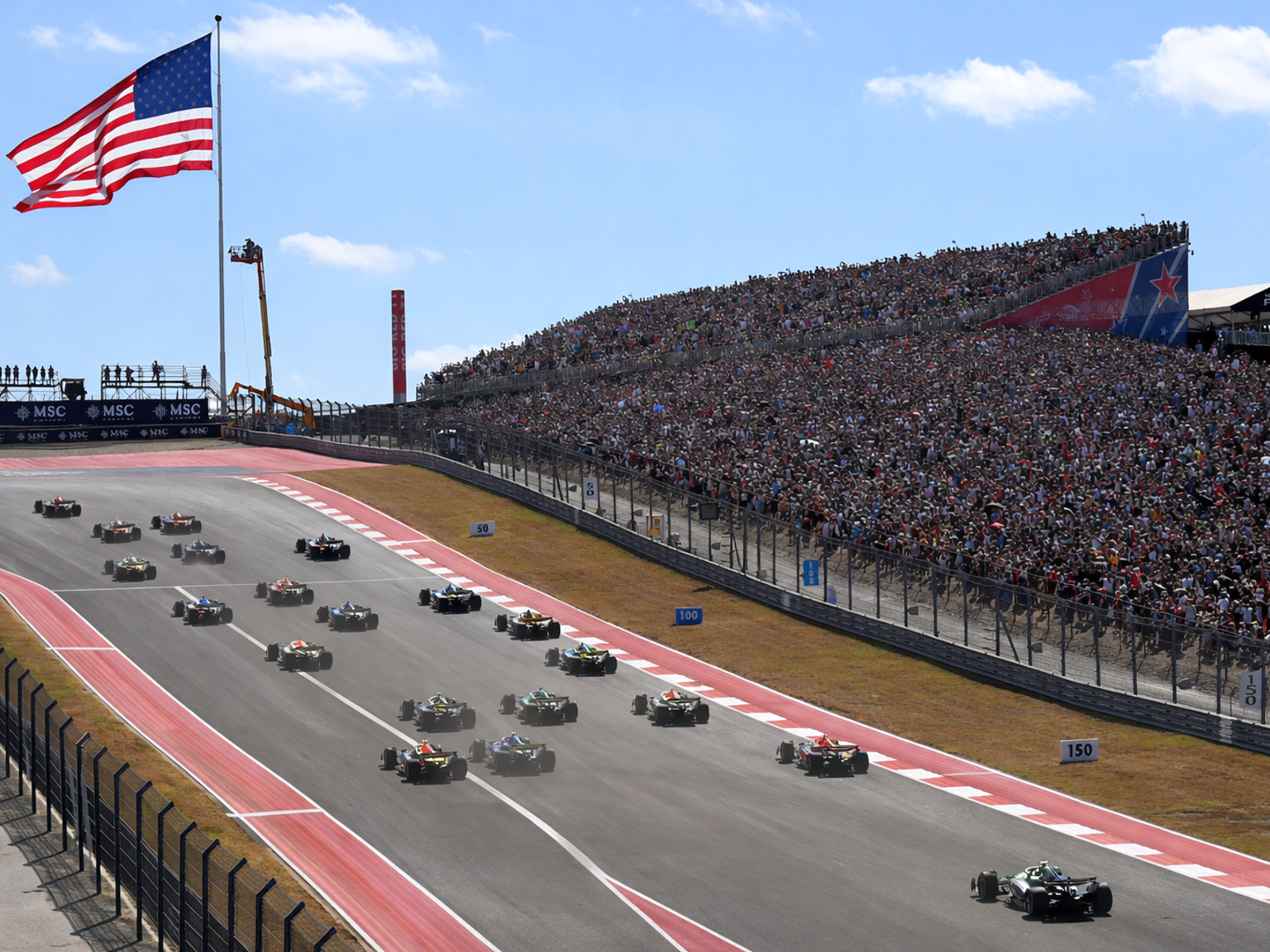 Open-wheel race cars on a track with a massive crowd in the grandstand, under a large American flag.
