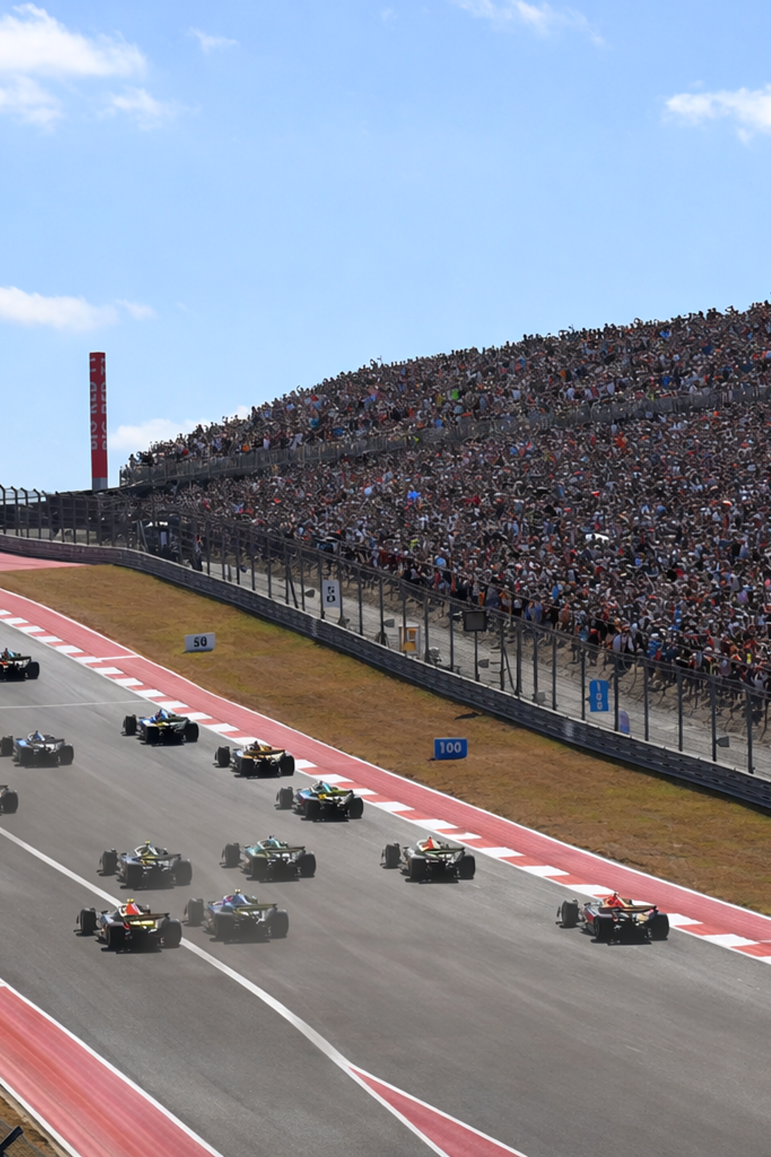 Open-wheel race cars on a track with a massive crowd in the grandstand, under a large American flag.