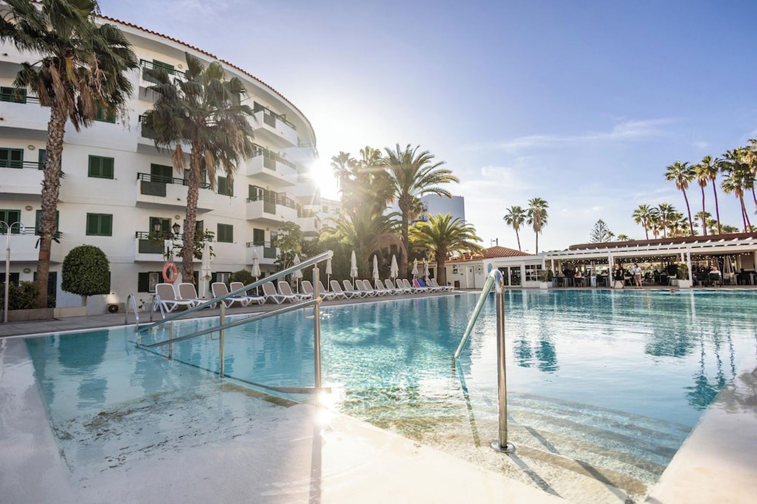 Utomhuspool på en solig resort med palmer och en vit byggnad.